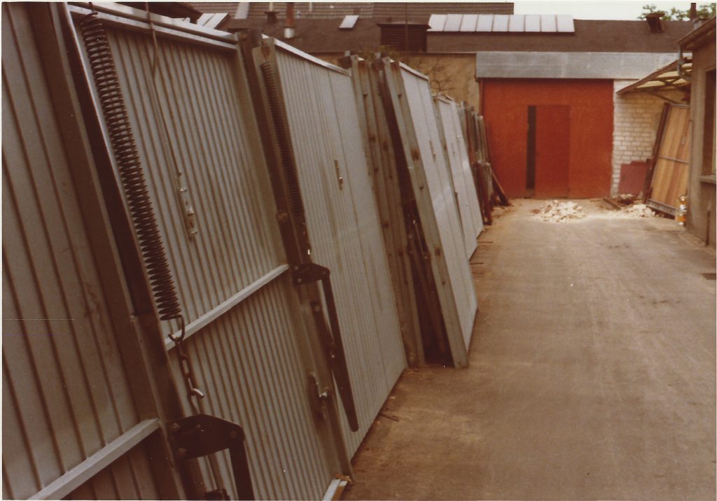 Älteres Foto: Mehrere Garagen-Schwingtore mit Federbeschlag lehnen in einem Hof an einer Wand.