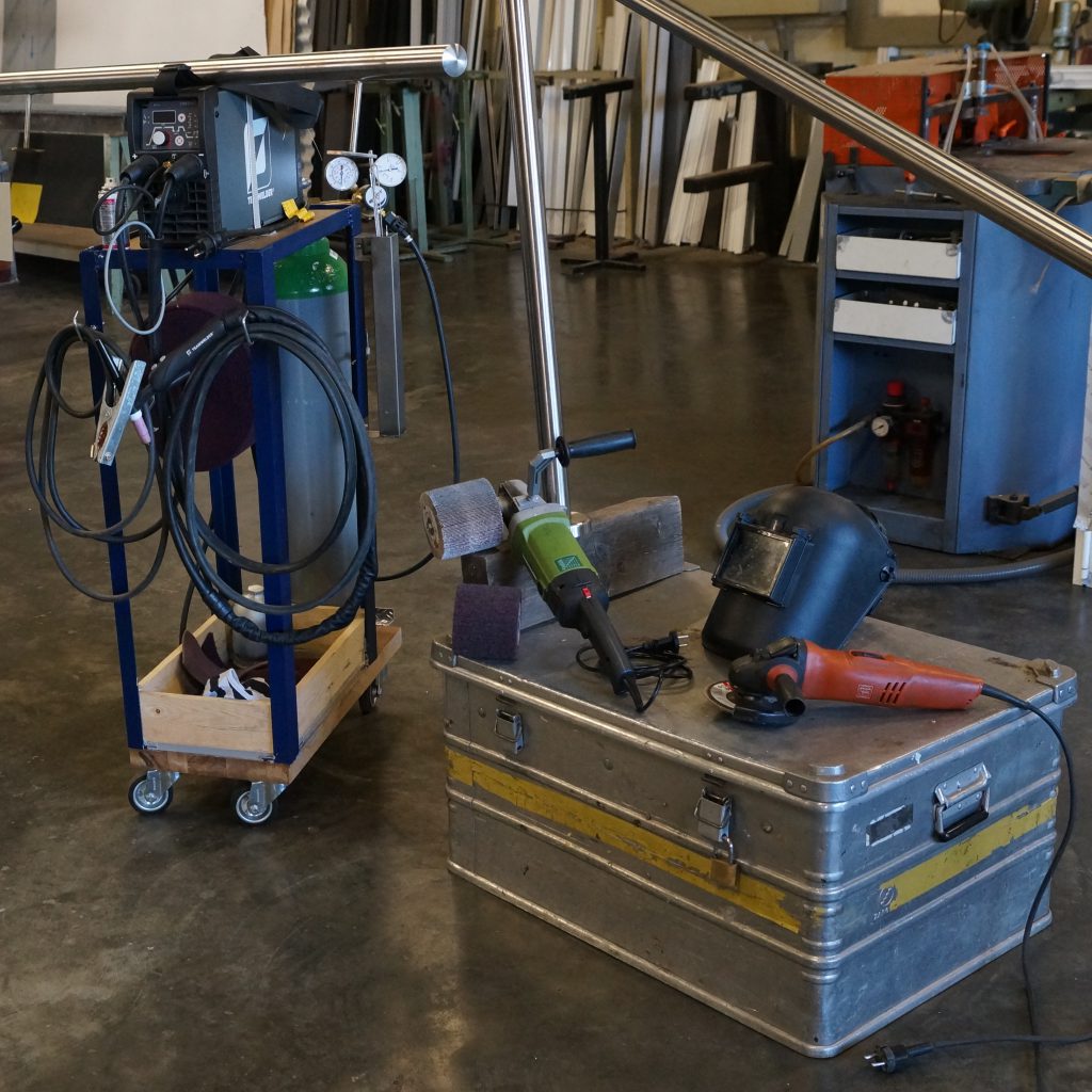 Schweißgerät auf einem Rollwagen mit Gasflasche, daneben eine Transportkiste mit Winkelschleifern und ein Schweißhelm; dahinter das Profillager.