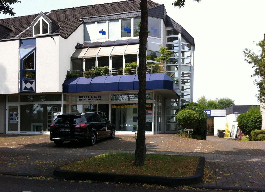 Das Betriebsgebäude an der Mielenforster Straße mit Markise und Schaufenstern, auf denen Wintergärten und Sonnenschutz stehen; vorn ein Baum.