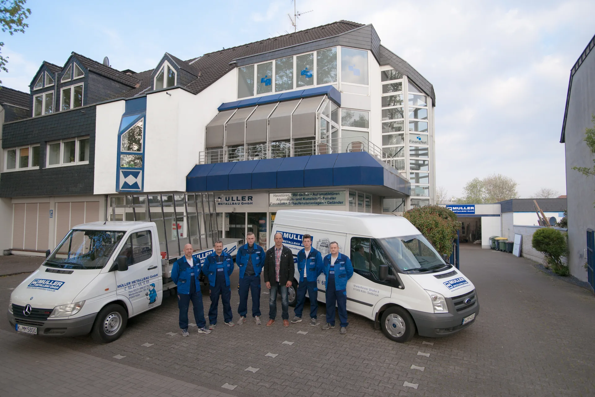 Sechs Männer, fünf davon in blauer Arbeitskleidung mit Firmenlogo, stehen vor dem Betriebsgebäude zwischen zwei weißen Transportern mit Firmenaufschrift.