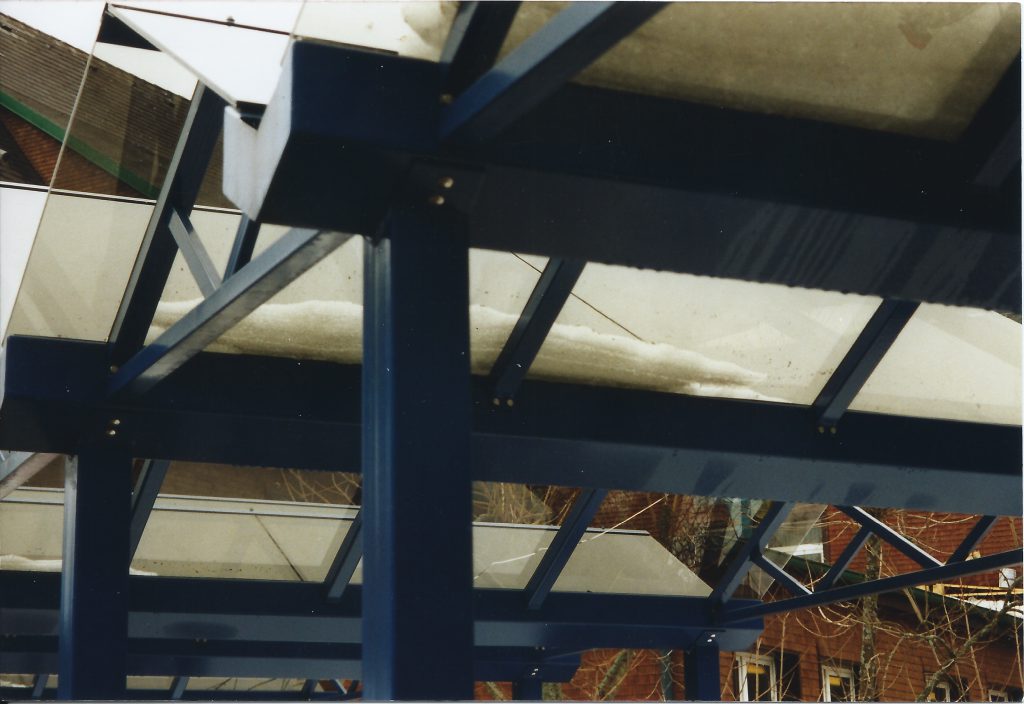 Älteres Foto: Untersicht einer blauen Profilkonstruktion mit Pfosten, Sparren und Streben unter einem Glasdach, dahinter ein Ziegelbau.