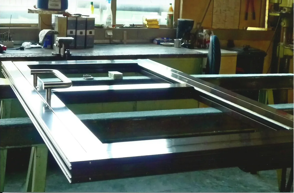 Ein dunkel beschichteter Türflügel mit langem Edelstahl-Stangengriff liegt auf Böcken in der Werkstatt, die Füllung ist noch offen.