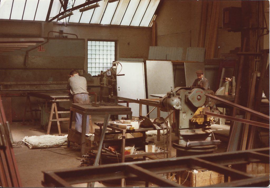 Historisches Foto der früheren Werkstatt mit Oberlichtern: zwei Männer an Werkbank und Maschinen, am Boden ein Stahlrahmen.